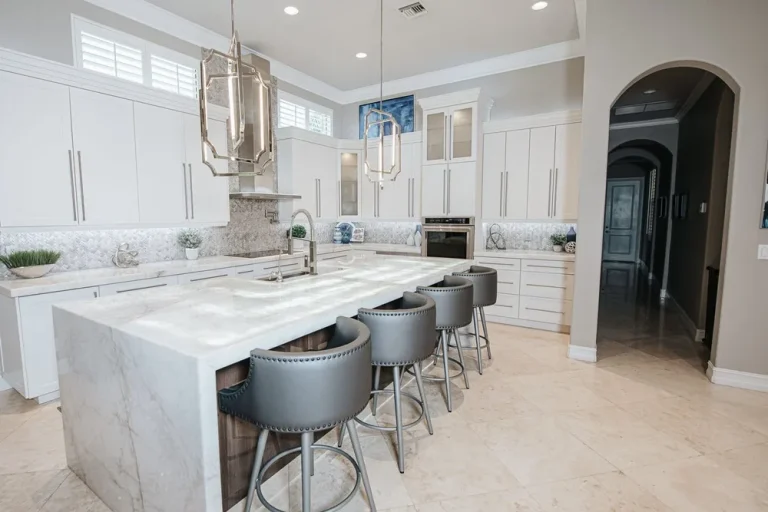 Modern white kitchen with marble island, gray leather bar stools, stainless steel pendant lights and beige tile flooring