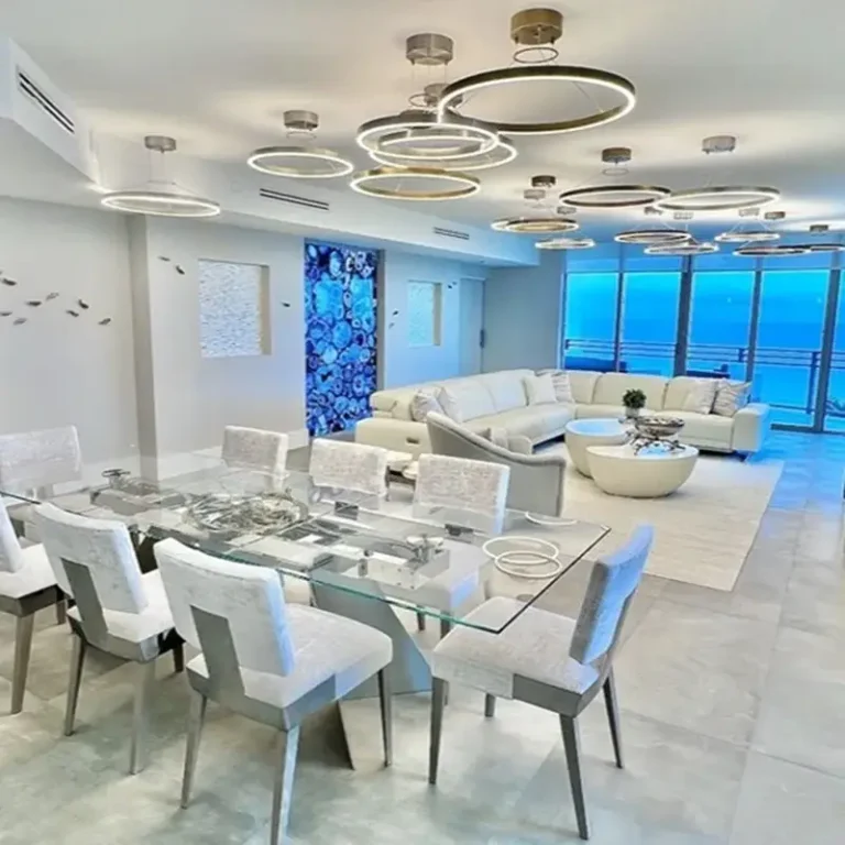 Modern luxury dining room with glass table, white leather chairs, circular pendant lights, and ocean view windows