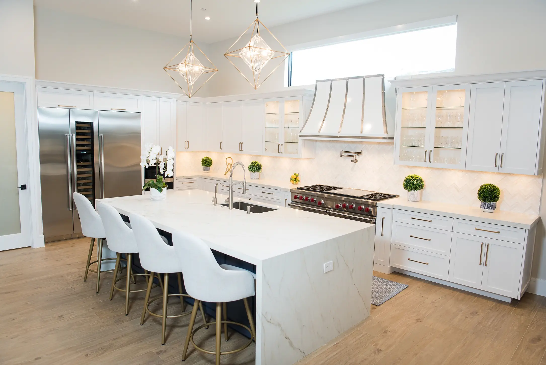 Modern white kitchen with quartz waterfall island, stainless steel appliances, geometric pendant lights, and white bar stools on light wood floors