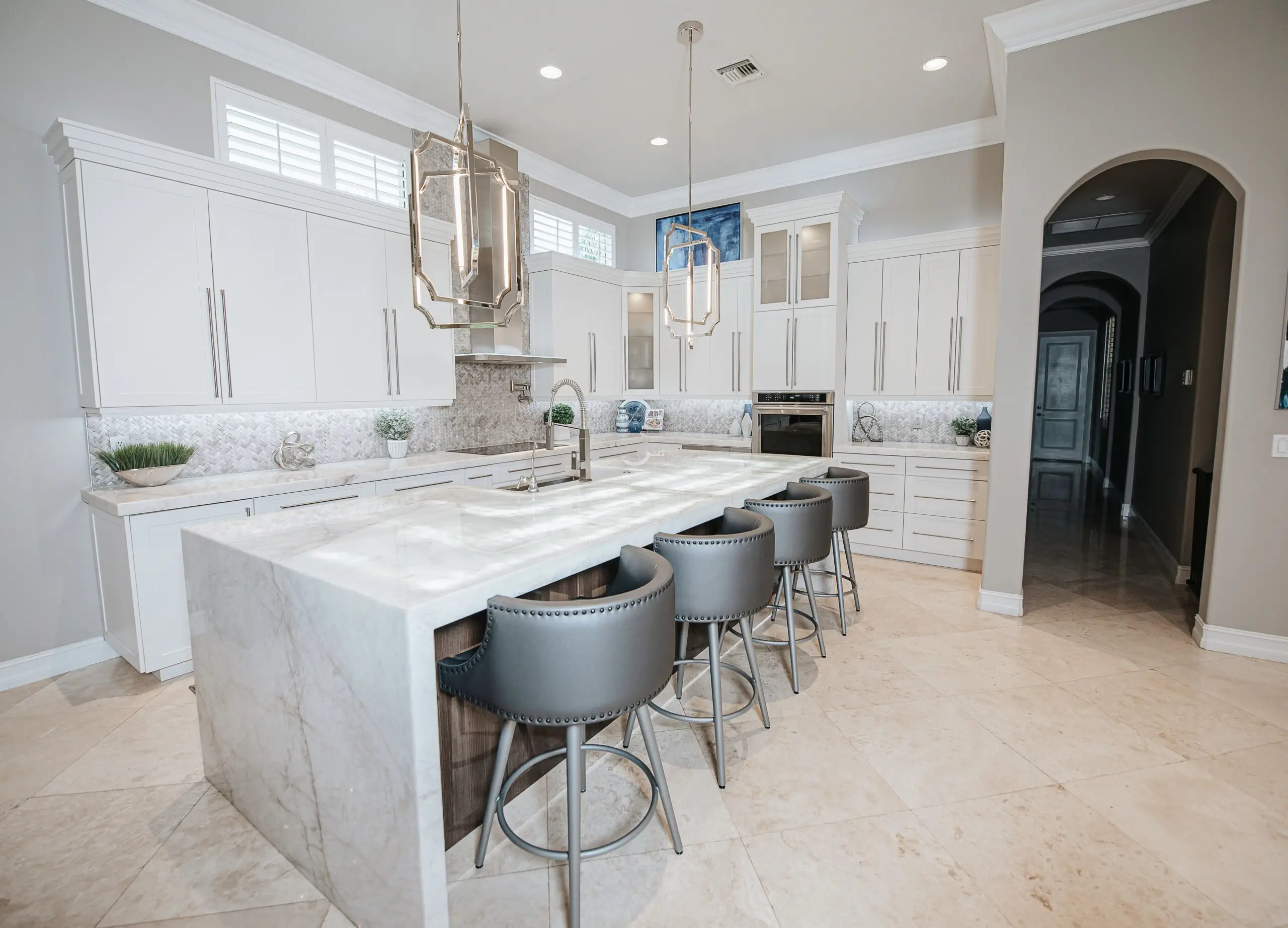 Modern white kitchen with marble countertops, pendant lighting, gray bar stools and travertine floors by YOLO Interiors Fort Lauderdale
