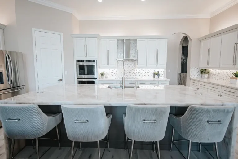 Modern white kitchen with marble countertops, herringbone backsplash, and light blue upholstered bar stools in luxury home