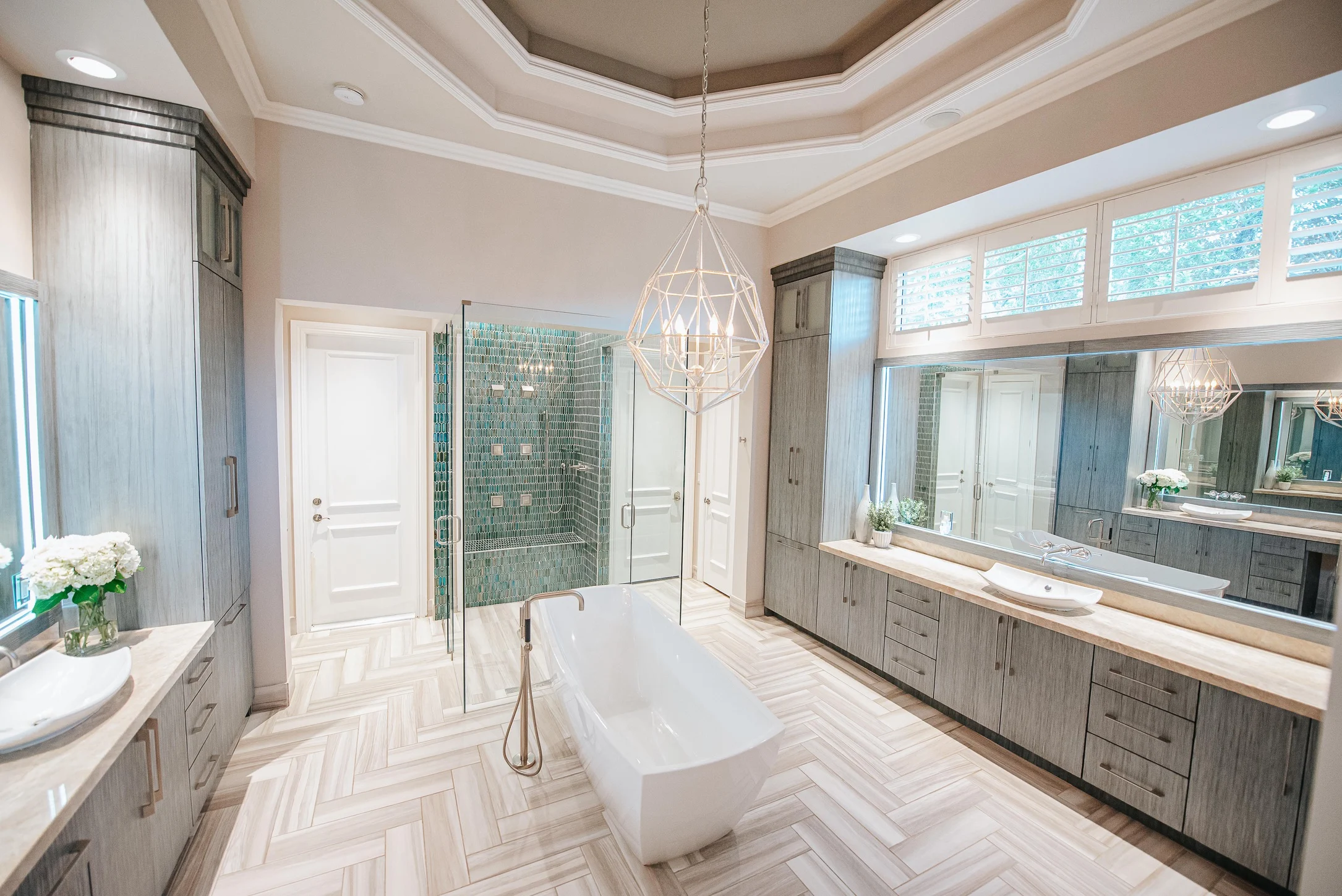 Luxury master bathroom with gray wood vanity, freestanding tub, geometric pendant lighting, teal glass tile shower, and herringbone wood floors