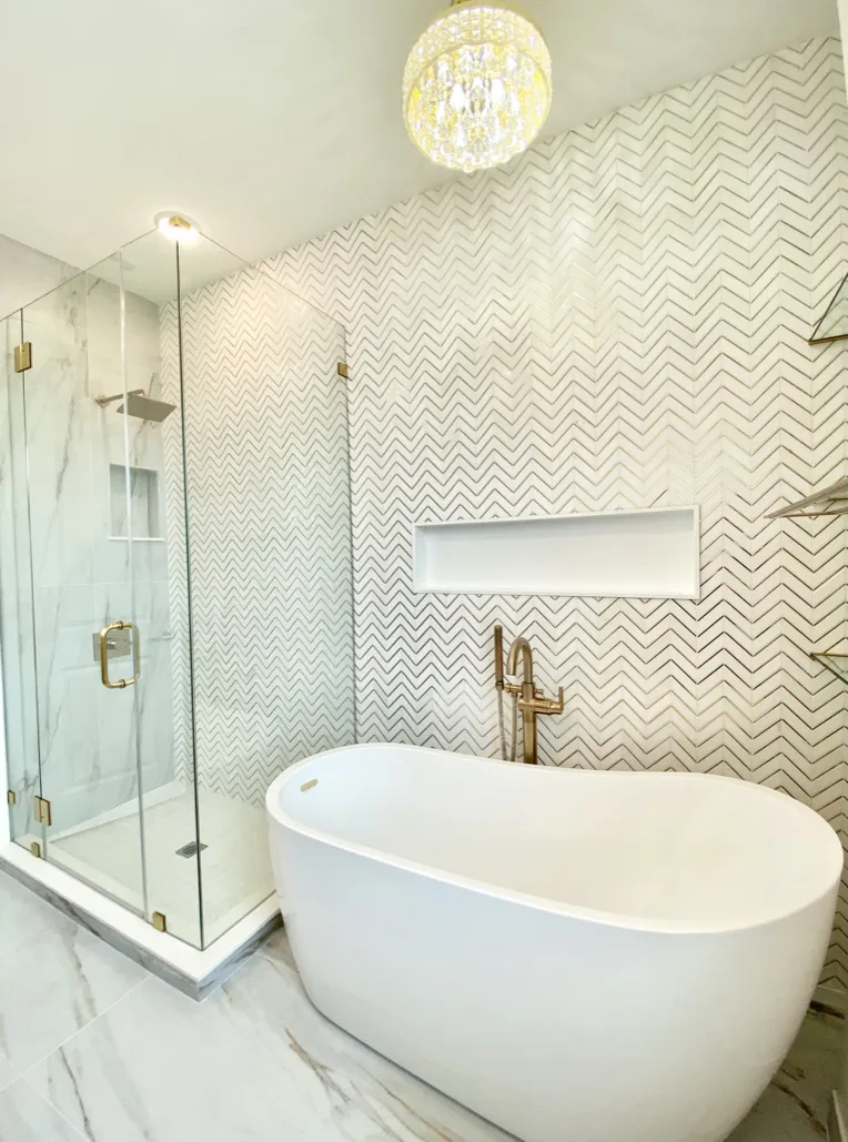Modern luxury bathroom with white freestanding soaking tub, chevron tile accent wall, glass shower enclosure, brass fixtures, and crystal chandelier in Fort Lauderdale