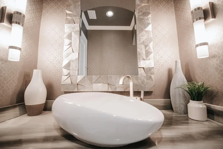 Modern luxury bathroom with white freestanding bathtub, geometric mirror, beige textured wallpaper, contemporary lighting fixtures and decorative vases