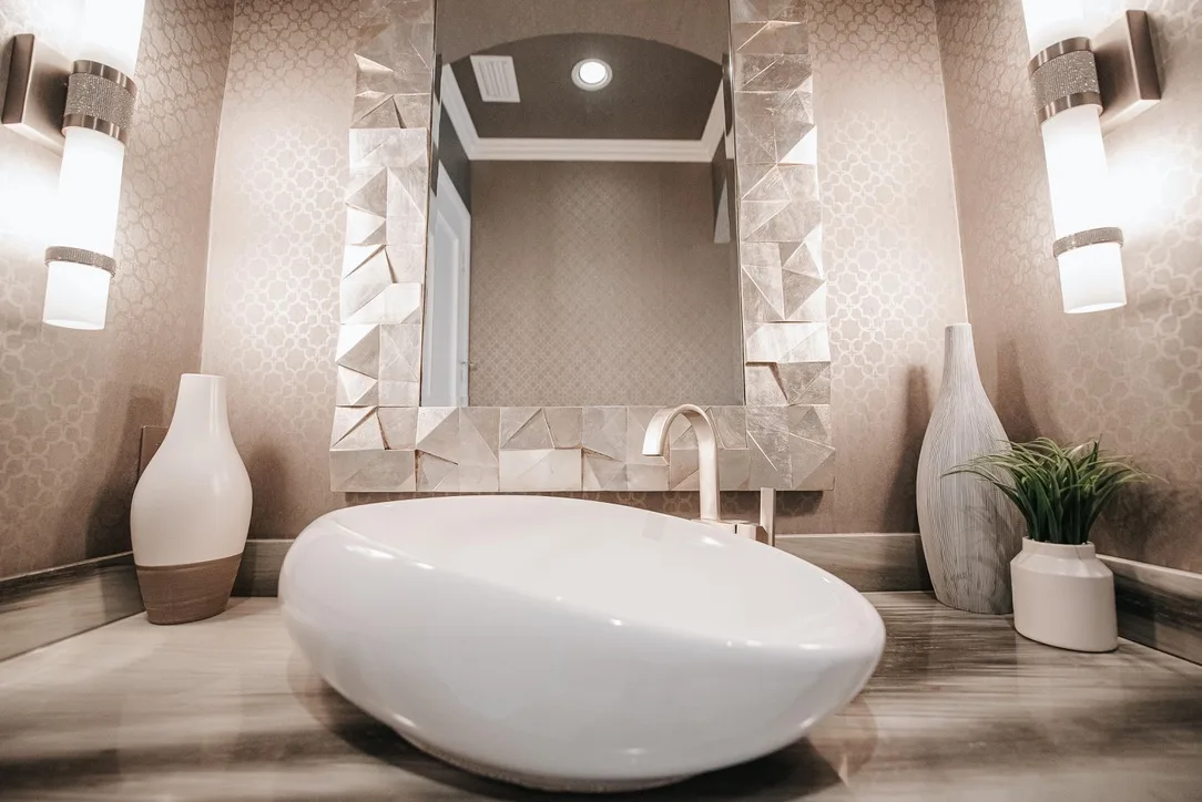 Modern luxury bathroom with white freestanding bathtub, geometric mirror, beige textured wallpaper, contemporary lighting fixtures and decorative vases