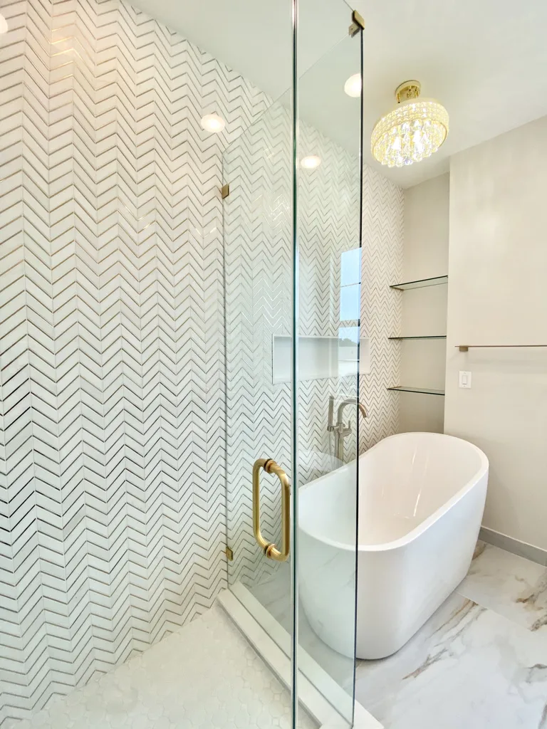 Luxury bathroom with white herringbone tile wall, freestanding white bathtub, glass shower door, brass fixtures, and crystal chandelier in modern interior design